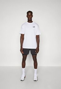 Balta T-veida krekls ar īsām piedurknēm un mazā melnā logo, kopā ar tumšām šortiem un baltām sporta kurpēm. Vienkāršs dizains, kokvilnas materiāls.