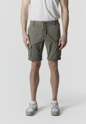 Uomo con bermuda cargo verde oliva con risvolti arrotolati, sneakers bianche e maglietta bianca, in piedi contro uno sfondo chiaro e uniforme.