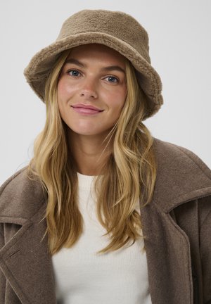 Brūns flīsa cepure ar mīkstu tekstūru, plašu malu un odere. Valkāta ar vieglu džemperi un brūnu puskombinezonu.