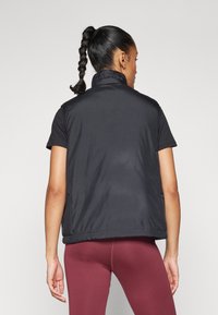 Svart ärmlös väst med ståkrage, slät textur och elastisk nederkant, buren över en åtsittande svart skjorta och vinröda leggings.