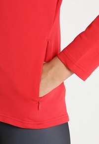 Červený top s dlouhým rukávem a zipovou boční kapsou, vyrobený z hladkého, elastického materiálu; má přiléhavý střih a viditelné švy.