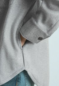 Gray wool blend shirt featuring a cuff with a button, side pocket, and rounded hem, complemented by a denim background.