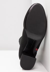 Semelle de bottine noire avec adhérence texturée, talon bloc, et détail subtil de rayure rouge, blanche et bleue sur le côté.