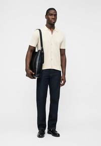 Άνδρας με κρεμ κοντομάνικο πουκάμισο με κουμπιά, σκούρο τζιν, μαύρα δερμάτινα παπούτσια, που κρατά μια μεγάλη μαύρη δερμάτινη τσάντα ώμου πάνω σε λευκό φόντο.