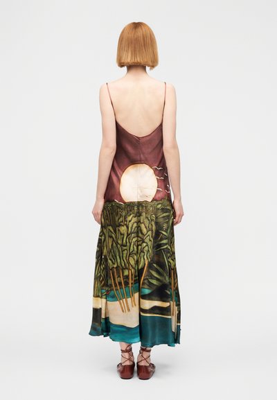 Γυναίκα με κοντά κόκκινα μαλλιά, που φοράει αμάνικο φόρεμα με σχέδιο ηλιοβασιλέματος, πουλιά και δασικό τοπίο, συνδυασμένο με καφέ δερμάτινα παπούτσια με κορδόνια.