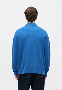 Homem com cabelo castanho encaracolado, vestindo um suéter azul largo e jeans escuros, de pé, de costas, contra um fundo claro e liso.