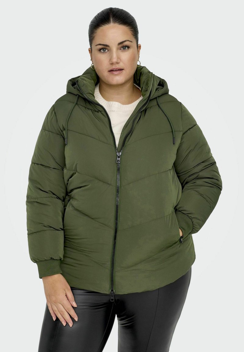 Žalias pūkuotas džemperis su kapišonu ir užtrauktuku, su zigzaguotomis juostelėmis ir tampriais rankogaliais. Dėvimas ant šviesios megztinio ir juodų kelnių.