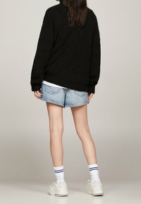 Kvinna som står med ryggen vänd, iförd en svart tröja, ljusa jeansshorts, vita strumpor med blå ränder och vita sneakers.