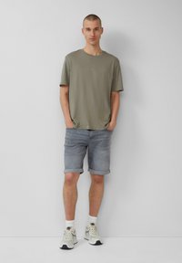 Licht olijfgroene t-shirt, korte mouwen, losse pasvorm. Lichtgrijze denim shorts, opgerolde mouwen. Witte en beige sportschoenen, enkelsokken.