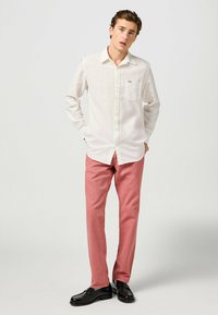 Camicia button-up beige chiaro con taschino sul petto, abbinata a pantaloni rosa chiaro e mocassini neri. La camicia ha maniche lunghe e colletto classico.