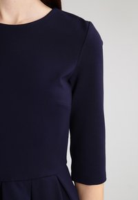 Námořnické modré šaty s kulatým výstřihem, tříčtvrtečními rukávy a rozšířenou sukní s hladkým povrchem. Minimalistický design.