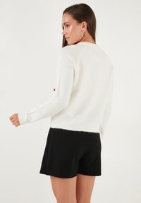 Witte gebreide trui met lange mouwen en geribbelde zoom, met een contrasterend rood detail op de mouw, gecombineerd met zwarte shorts.