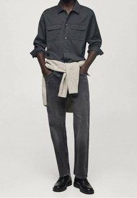 Camicia grigia scura con bottoni e due tasche sul petto, abbinata a jeans grigi sbiaditi. Maglione annodato in vita, scarpe di pelle nere.