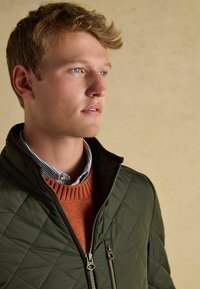 Mladý muž s modrými očami v zelenej prešívanej bunde, oranžovom svetri a modrej pruhovanej košeli, pozerajúci sa do strany na béžovom pozadí.