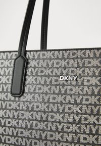 Μαύρη και γκρί τσάντα tote με λογότυπο DKNY, διαθέτοντας υφασμάτινη επιφάνεια με επαναλαμβανόμενα γράμματα και κομψές δερμάτινες λαβές.