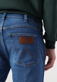 Los jeans de mezclilla azul cuentan con una etiqueta de cuero marrón con el logotipo "Wrangler", dos bolsillos traseros con costuras en contraste y un diseño ajustado.