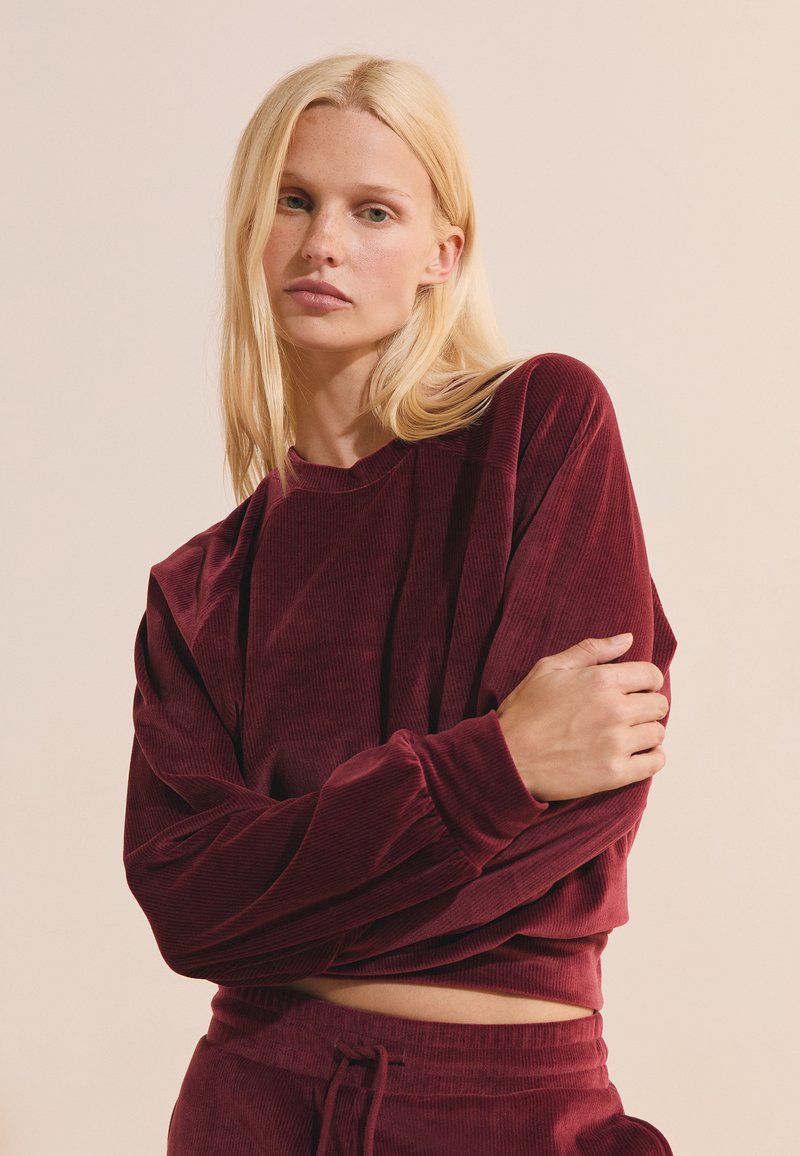 Mujer rubia de pelo liso con sudadera burdeos de pana de manga larga y pantalones a juego, posando con un brazo cruzado sobre el cuerpo.