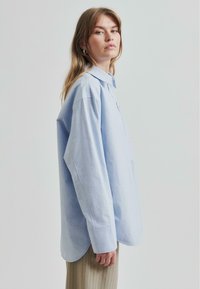 Cămașă oversized de un albastru deschis, confecționată dintr-un material neted, cu guler, mâneci lungi și tiv curbat. Stilizată cu pantaloni cu nervuri de culoare deschisă.