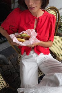 Rote Bluse mit floralen Stickereien, weiße gestreifte Hose, hält ein in pinkem Papier eingewickeltes Sandwich mit Salat und einem knusprigen frittierten Patty.