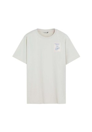 Camiseta de manga corta de color beige claro con cuello redondo y un pequeño parche rectangular en el pecho izquierdo con un diseño náutico y texto.