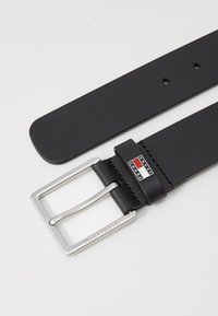 Zwarte synthetische riem met een zilveren rechthoekige gesp. Heeft een logo label en een gestikte rand. Gladde textuur, eenvoudig ontwerp.