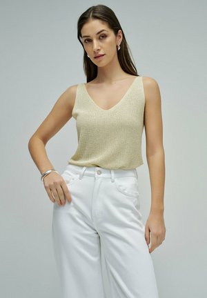 Joven mujer que lleva una camiseta sin mangas de punto beige y pantalones blancos de talle alto, posando con una mano en la cadera contra un fondo liso.