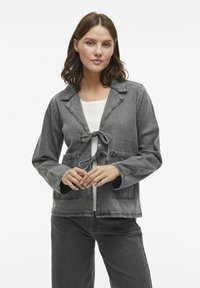 Sivá deninová bunda s golierom, viazaním na šnúrku a prednými vreckami. Má uvoľnený strih a dlhé rukávy, skombinovaná s svetlou blúzkou.