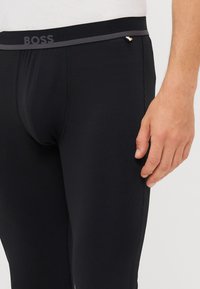 Svarta leggings med en åtsittande design, featuring ett kontrasterande grått elastiskt midjeband med "BOSS"-logotyp, slät textur och inga synliga mönster.