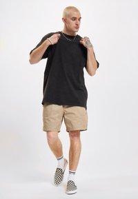 Tricou oversized negru, pantaloni scurți bej, șosete albe cu dungi negre și pantofi slip-on în carouri alb-negru. Modelul pozează central.