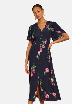 Žena má na sebe námorníckomodré midi šaty s ružovým kvetinovým vzorom, čipkovaný výstrih do V, krátke nabíjané rukávy, gombíky vpredu a predný rozparok.