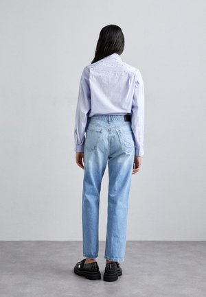 Kobieta o długich ciemnych włosach stoi zwrócona twarzą do szarej ściany, ubrana w jasnoniebieską koszulę w kratę, niebieskie dżinsy z wysokim stanem oraz czarne buty.