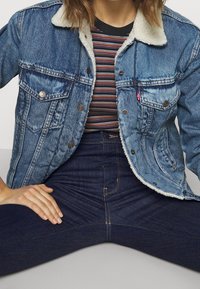 Chaqueta de mezclilla con un lavado azul claro, cuello de borreguito, dos bolsillos en el pecho y botones plateados, llevada sobre una camiseta de rayas y jeans oscuros.