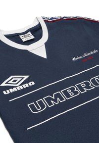 Tummansininen urheilupaita, jossa on valkoinen kaula-aukko ja yksityiskohtia, etupuolella valkoinen "UMBRO"-logo ja punaisella brodeerattu "Umbro Manchester".