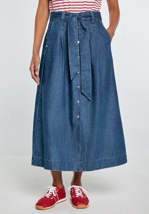 Kobieta nosząca niebieską jeansową spódnicę do kostek z guzikami z przodu i wiązaniem w talii, połączoną z czerwonymi trampkami i pasiastym topem.