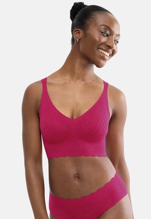 Femme souriante avec les cheveux attachés en chignon, portant un soutien-gorge sans couture magenta assorti et une culotte taille haute avec un motif texturé subtil.