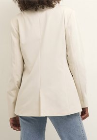 Femme aux cheveux bouclés portant un blazer crème et un jean bleu, vue de dos sur un fond clair uni.