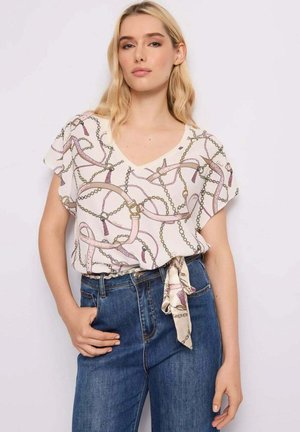 Mujer rubia con blusa blanca de escote en V con estampado de cinturón rosa y cadena, anudada en la cintura, combinada con vaqueros azules de talle alto.