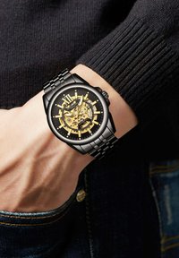 Zwarte polshorloge met een skelet wijzerplaat en gouden accenten. Heeft grote Romeinse cijfers en een band van roestvrij staal. Textuur van een zwarte trui als achtergrond.