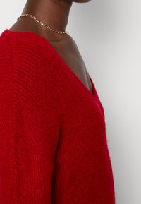 Pull en tricot rouge avec un décolleté en V profond et un tissu texturé. Un collier en chaîne dorée porté autour du cou ajoute une touche métallique.