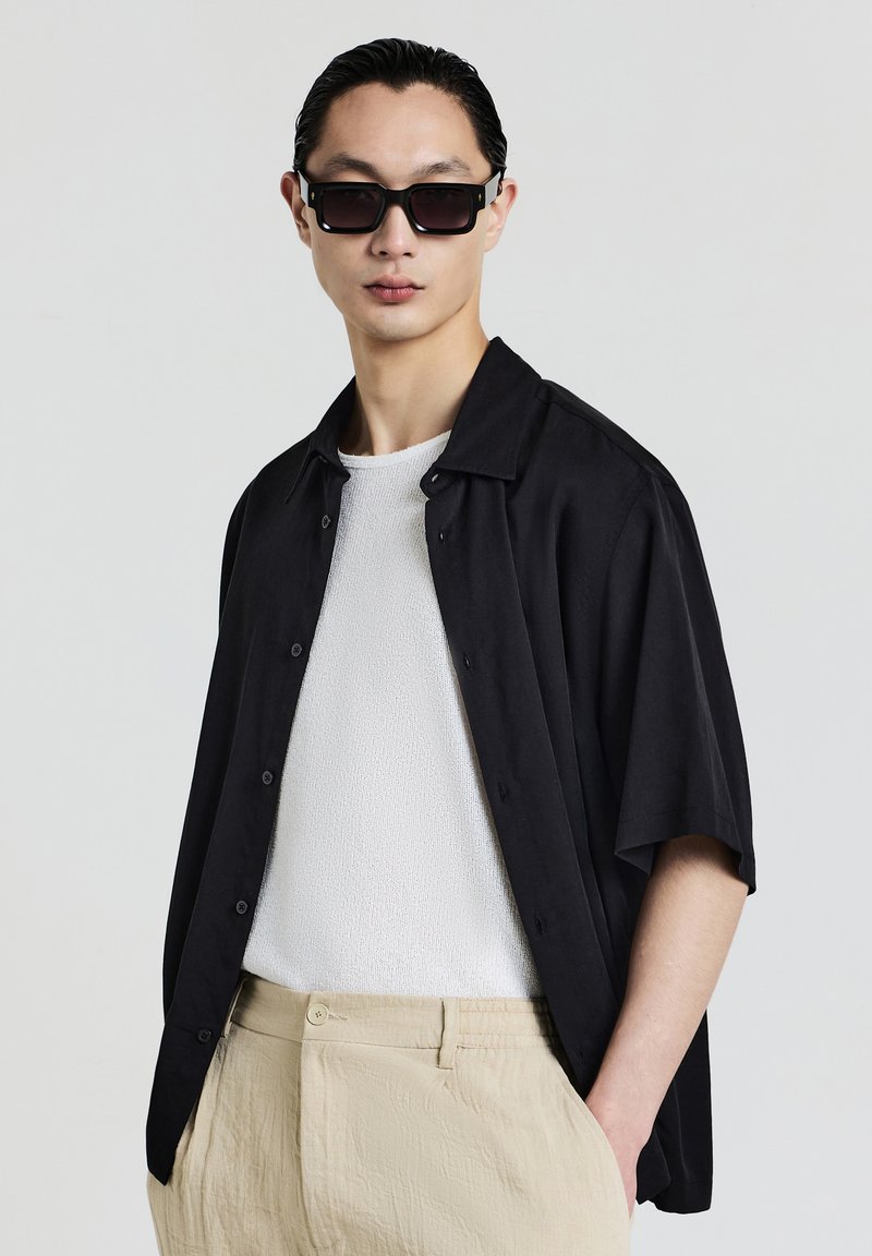 Uomo che indossa occhiali da sole neri oversize, una camicia nera a maniche corte aperta sopra una camicia bianca con texture e pantaloni beige, con le mani nelle tasche.