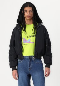 Black puffer jacket with a hood, featuring a zip closure and side pockets. Paired with a bright lime green graphic t-shirt and blue jeans.