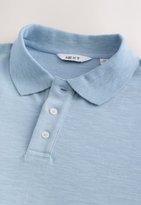 Polo bleu clair avec un col à revers, trois boutons blancs, et un tissu texturé. Étiquette de la marque visible au niveau du col.