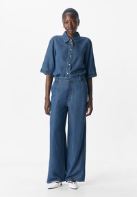 Femme portant une chemise en denim bleu ample avec des boutons et un pantalon en jean large assorti, associée à des escarpins blancs à bout pointu.