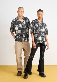 Camicie a righe bianco e nero indossate da due persone, con maniche corte e vestibilità ampia. Pantaloni color sabbia chiara e pantaloni neri svasati completano gli outfit.