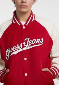 Crvena jakna u varsity stilu s kremastim rukavima, rebrastim ovratnikom i bijelim dugmadi za snap. Logo "Guess Jeans" u bijelom izvezenom slovu.