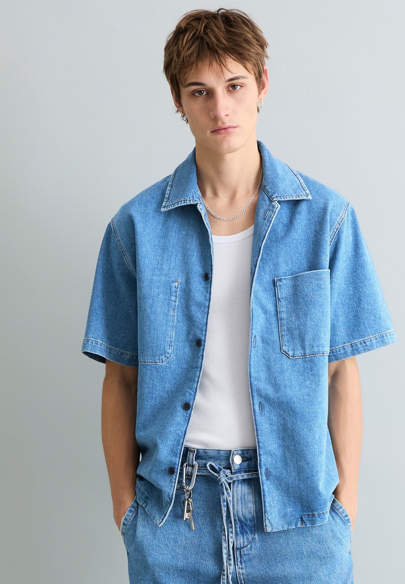 Jeune homme portant une chemise en jean à manches courtes bleu clair par-dessus un débardeur blanc, un pantalon en jean assorti, un collier chaîne en argent, et un porte-clés accroché à la boucle de sa ceinture.