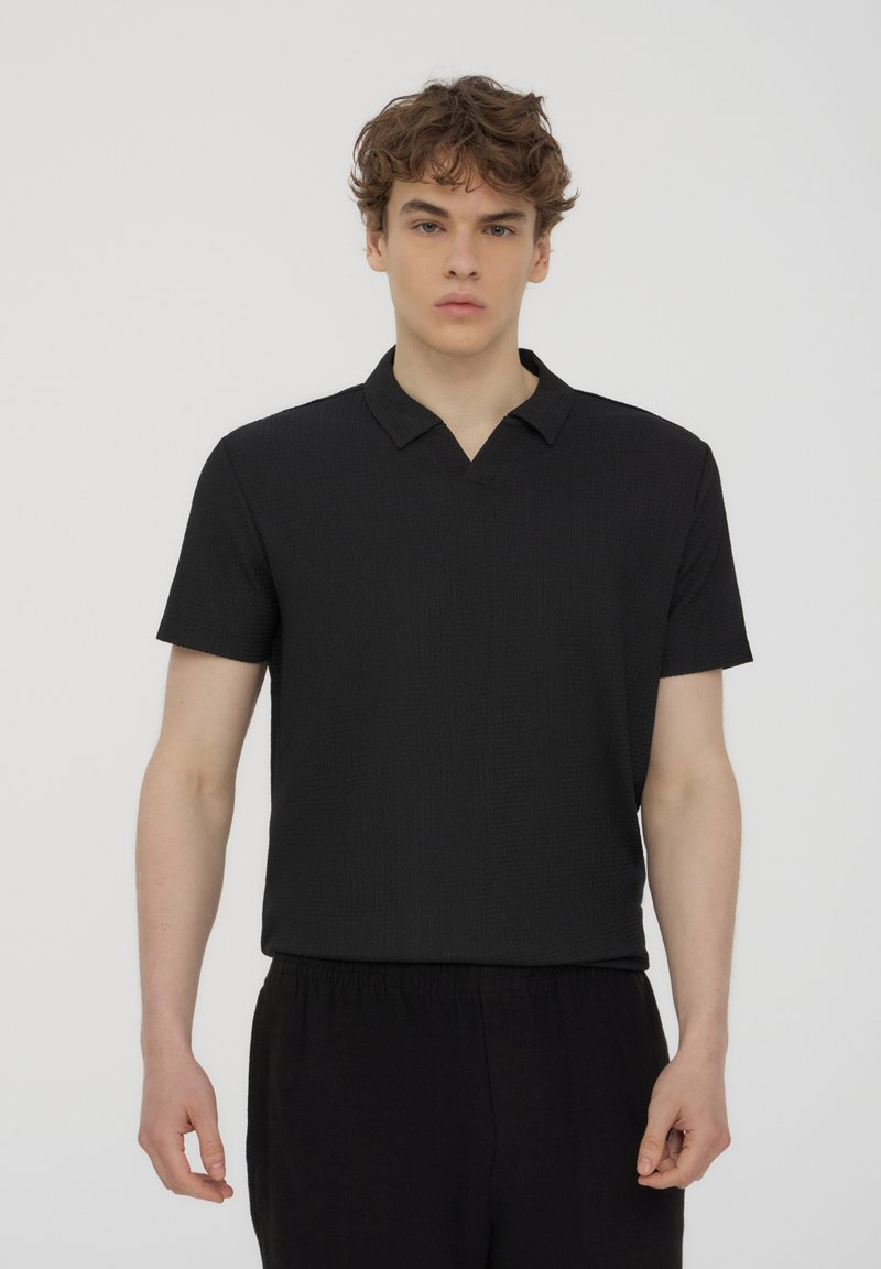 Polo shirt nero a maniche corte con tessuto texturizzato, caratterizzato da un design a V e una vestibilità casual. Indossato con pantaloni neri larghi.