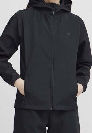 Person iført en sort hættetrøje med hænder i lynlåslommerne på siderne, stående mod en ensfarvet lys baggrund.