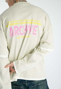 Beige Langarmhemd mit pink und gelbem grafischen Text auf dem Rücken. Mit weißen Streifen an den Ärmeln und einem lockeren Schnitt.