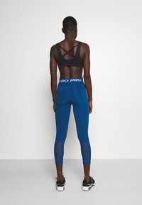 Person som står vänd bort med svart sportbh och blå högmidjade leggings med "PRO" midjeband, samt svarta träningsskor.